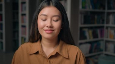Yakın plan mutlu Asyalı kadın kütüphanede poz veriyor. Masasında oturan fotoğrafçı kız öğrenciye bakıyor. Üniversite sınavına hazırlanıyor.