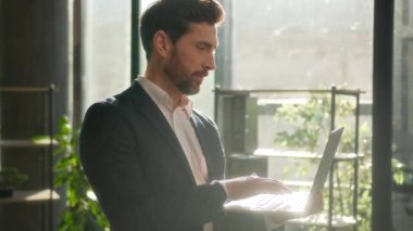 Caucasian businessman typing on laptop checking email standing in office morning in sunbeams sunrise man successful entrepreneur professional manager leader corporate executive smiling looking camera