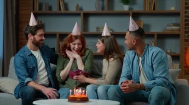 Excited woman receive gift present from friends diverse multiracial colleagues congratulate wish happy birthday presenting surprise box. Ethnic women men in party caps have fun celebrate event at home