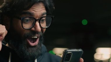 Gözlüklü mutlu, sakallı Hintli iş adamı otoparktaki akıllı telefondan iyi haberler alıyor. Heyecanlı erkek kazanan Arap adam zaferi cep telefonuyla kazandı.