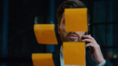 Caucasian businessman marketing manager middle-aged 40s man talk mobile phone multitask working in night office write sticky notes on glass wall data market analysis research business planning project