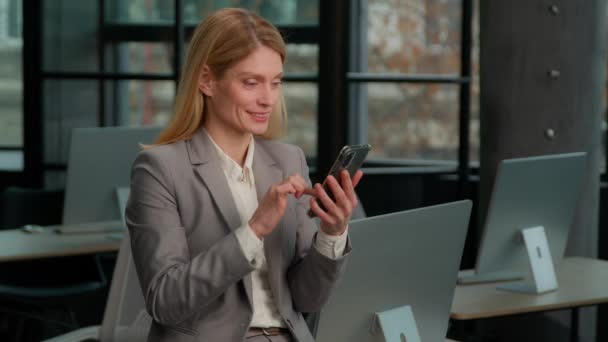 白种人中年妇女微笑着拿着智能手机刷页在应用社交网络应用选择商品站在办公室的网店 有手机浏览业务的成年女商人 — 图库视频影像