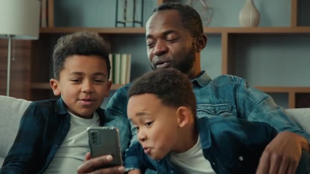 アフリカ系アメリカ人の家族の父男と小さな子供男の子子供はソファの上に座っている携帯電話を使用して楽しいを持っている携帯電話は面白いアプリの写真マスクを試してみてください自己愛の顔をした携帯電話で家を見て — ストック動画