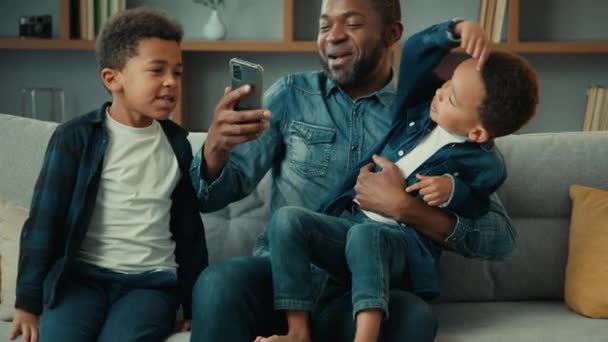 快乐的非洲裔美国人家庭客厅里的成人父母爸爸爸爸爸爸爸爸看手机坐在沙发上和可爱的男孩孩子们儿子们拥抱在沙发上笑着开心地笑着 — 图库视频影像