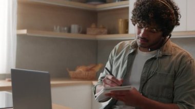 Kulaklıklı Hintli bir adam yurt içi mutfağında oturmuş laptopla ilgili eğitim videosu dersi izliyor. Arap öğrenci uzak online karantina dersleri alıyor.