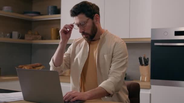 キッチンでノートパソコンを使用している白人男性はメガネの眼の疲れに苦しんでいます 疲れた男性フリーランスのビジネスマン自宅でコンピュータと仕事眼鏡疲れ視力と目の痛み不快感 — ストック動画