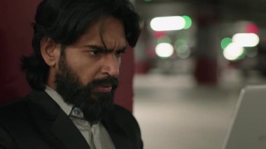 Close-up puzzled serious Indian man looking at laptop screen in underground parking focused worried young arabian businessman feeling anxiety from reading bad business news working remote on Internet