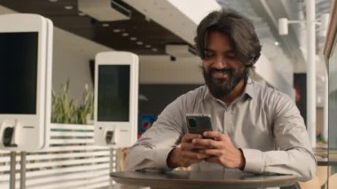 Happy young indian businessman holding smartphone sending message sitting indoors smiling arabian man play mobile game using application on phone reading good news on internet chatting in social media