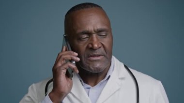 Olgun Afro-Amerikan erkek doktor telefonda, beyaz önlüklü, kafası karışık bir erkek tıp danışmanıyla konuşuyor. Akıllı telefon müşterilerine danışıyor.