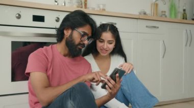 Arap Hintli ırklar arası kadın erkek, mutfakta sıcak bir zeminde oturup sosyal medyada komik videolar izleyip çevrimiçi konforlu dağıtım hizmetleri sipariş ediyor. Cep telefonlarını internet üzerinden satın alıyorlar.