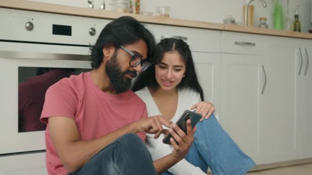アラビア語インドの異人種間のカップルの女性の男性は オンライン快適配信サービスを注文ソーシャルメディアで面白いビデオコンテンツを見てキッチンで暖かい床に座っているインターネット上で携帯電話の購入商品を保持 — ストック動画
