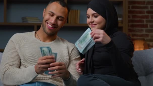 多様な民族のカップルがソファに座ってお金を数えるドル紙幣アフリカ系アメリカ人男性の夫は ヒジャーブ計画のイスラム教徒の妻に財政の一部を与える家族の予算豊かなパートナーは現金通貨を数える — ストック動画