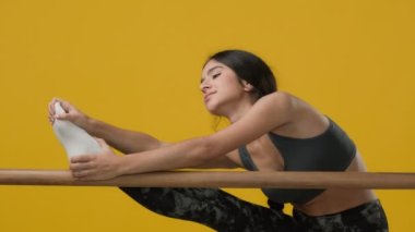 Üzgün etnik Hintli genç, esnek kadın balerin akrobat bayan akrobat kız, stüdyo sarı arka planda bale çubuğuyla bacaklarını esnetiyor zor güç güç gerilim koreografisi
