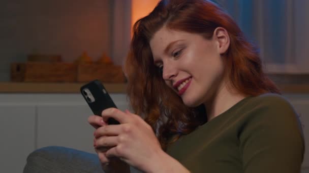 幸せなリラックスしたCaucasian Redhead女性自宅でソファの上で自宅で電話でソーシャルネットワークでチャットスマートフォンの画面を見て笑顔の女の子は 携帯電話を使用してインターネット上でモバイルショッピングアプリを楽しんで — ストック動画
