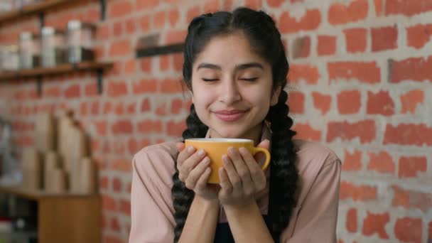 年轻快乐的阿拉伯女人在咖啡馆里享受着芬芳的饮料 面带微笑轻松地印度女孩餐厅的顾客在自助餐厅里享用着美味的拿铁卡布奇诺呼吸着新鲜的咖啡 看着相机 — 图库视频影像