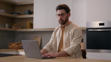 40 'lı yaşlarda ciddi bir beyaz erkek ev sahibi erkek bilgisayarını mutfakta gezdirip kameraya bakarak internetten alışveriş yapıyor. Masada bilgisayar servisi pozu veren serbest çalışan bir iş adamı.