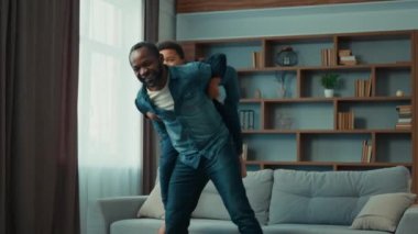 Mutlu bir aile babası Afrikalı bir baba sırtına binmiş komik bir oğlan çocuğu ile dans ediyor evde neşeli bir şekilde dans ediyor rahat oturma odasında çocuk bakımında birlikte oynuyorlar.