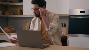 Beyaz bir adam ev sahibi, evrakları kontrol ediyor. Finansal evrakları inceliyor. Mutfakta dizüstü bilgisayarla ev kredisi ödemesi yapıyor. Evde çalışan serbest çalışan işadamı.