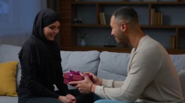 Çok ırklı çift, evlilik yıldönümünü kutluyor. Afrikalı Amerikalı erkek, tesettürlü Müslüman kadına hediye veriyor. Uluslararası kadınlar için sürpriz hediye paketi.