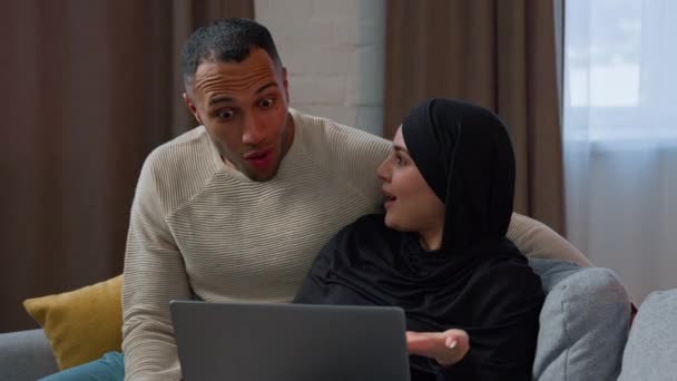 夫婦はノートパソコンの画面の話を見ます承認銀行のクレジットを受け取ります幸せな多人種間のアフリカ系アメリカ人男性とイスラム教徒の女性でヒジャーブ勝利オンラインベット宝くじ勝利インターネットショッピング自宅で — ストック動画