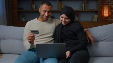 Çok ırklı gülümseyen bir çift evde dizüstü bilgisayar siparişi kullanırken kredi kartı kullanan tesettürlü Müslüman kadın ve Afrikalı Amerikalı koca müşterileri online alışveriş dijital ödeme e-bankasına ödeme yapıyorlar.