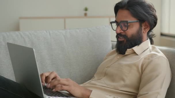 アラビア語インドのイスラム教徒の男男性ビジネスマンフリーランスソファの上に横たわっ仕事リラックス自宅タイピングラップトップキーボードのWebサーフィンチャットレポートの話ビジネスパートナーとオンライン話を書く — ストック動画
