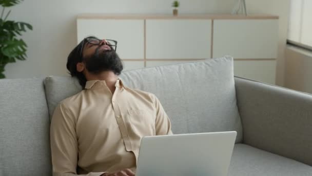 疲れきったフリーランスは自宅でノートパソコンで作業過労アラブのイスラム教徒の男はソファの上で眠りに落ちる疲れた睡眠インドのビジネスマンは ソファの疲労疲労にもたれる休憩休憩休息リラックスが必要 — ストック動画