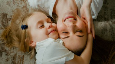 Cute affectionate adopted little kid preschool girl hugging cuddling mother head top view. Child daughter with mum two faces cuddle lying down at home talking smiling with eyes closed tender moment