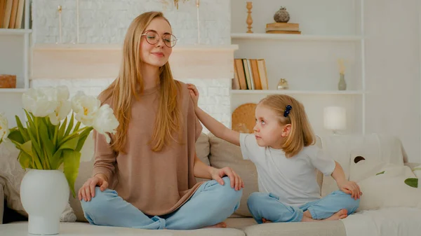Mindful Caucasian mother calm woman meditating with closed eyes in lotus position doing yoga exercise meditation sit on sofa at home little child girl daughter shake mom need attention distract to mum