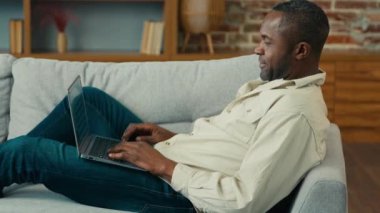 Afro-Amerikalı bir adam oturma odasında rahat bir şekilde yatıp dizüstü bilgisayarda yazı yazıyor. Yetişkin bir etnik işadamı. Evde internet üzerinde çalışıyor.