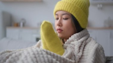 Sıcak şapkalı ve eldivenli hasta Asyalı kadın soğuk mutfakta oturuyor. Donmuş, çok ırklı Koreli Japon kız battaniyeye sarılı. Soğuk algınlığı var.