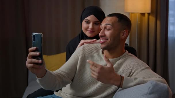 多民族カップルアフリカ系アメリカ人男性とイスラム教徒の女性でヒジャーブはソファの上に座っている携帯電話を使用してモバイルビデオチャットアプリ遠くの呼び出し多民族の夫の妻は ウェブカメラのスマートフォンの話をオンラインで話す — ストック動画
