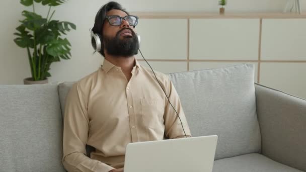 疲惫的穆斯林男子穿着耳机在家休息笔记本电脑工作 听着轻松的音乐 精疲力竭的商人头戴耳机放松听着和平旋律歌曲和平宁静安详的压力自由自在地躺在沙发上 — 图库视频影像