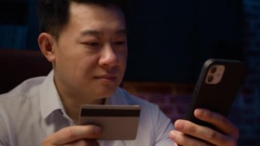 Karanlık bir gecede gülümseyen mutlu Asyalı adam cep telefonuyla kredi kartı kullanan Koreli erkek işadamı kolay internet üzerinden cep telefonu alışverişi indirimi ile ödeme rezervasyonu yaptırıyor.