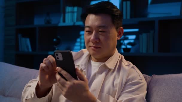 アジア民族の男夜の家でソファスクロール携帯電話のソーシャルメディアブラウジングインターネットサービスオンラインスマートフォンで買い物韓国の大人の男性と電話でデートアプリを使用して笑顔 — ストック動画