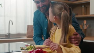 Sevimli küçük beyaz kız çocuk sebze salatası ve sebze karışımı yapıyor. Mutfakta sağlıklı vejetaryen yemekleri pişiriyor. Babasına sarılıyor, kızına sarılıyor.