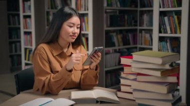 Üniversite kütüphanesindeki gülümseyen Asyalı kadın öğrenci kontrol zaman çizelgesi cep telefonu bilgilerinde sosyal medyada sohbet eden Çinli bir kıza cep telefonu uygulaması kullanarak öğrenmek için ara verin