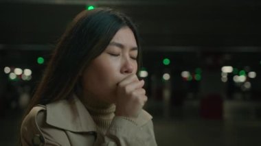 Sağlıksız hasta Asyalı kadın grip belirtilerine karşı öksürüyor Çinli Japon kadın soğuk, karanlık bir otoparkta, boğazı ağrıyan, öksürük şurubu enfeksiyonu olan Koreli kadının yanında duruyor.