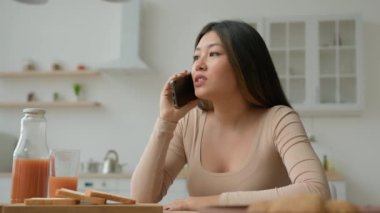 Asyalı kadın, ev mutfağında cep telefonuyla konuşuyor. Çok ırklı Çinli kız arkadaşlarıyla sohbet ediyor. Gülümsüyor, telefonla konuşuyor.