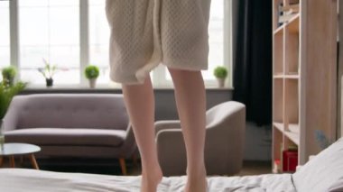 Hareketli kadın bacakları yatağa atlıyor Kafkasyalı mutlu kadın kaygısız ev sahibi kadın bornoz içinde yatak odasında zıplarken eğleniyor hafta sonu otel tatilinde eğleniyor.