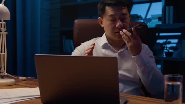 夜のアジアの中年男性のオフィス労働者ノートパソコンの作業と韓国の男性ビジネスマンの幹部は 音声Aiスマートフォン技術と電話での回答にビジネスオーディオメッセージボイスメールを指示します — ストック動画