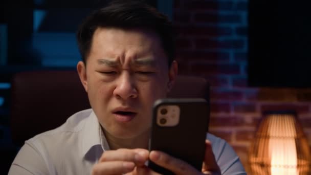 夜のアジア系成人男性自宅で夜のオフィスで失敗を失った携帯電話悲しい不幸な中国人男性スマートフォンで悪いニュースを失う不満韓国の中年ビジネスマンの携帯電話の問題 — ストック動画