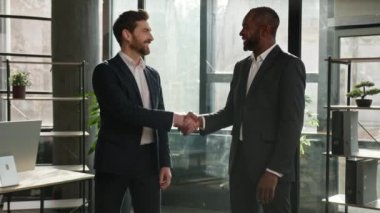 İki farklı etnik çeşitlilikte iş adamı iş ortağı. Ofiste kameraya gülümseyen adamlar ve Afrikalı Amerikalı çalışanlar el sıkışırken ortaklık anlaşması imzalıyorlar.