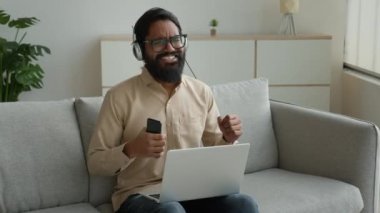 Kulaklıklı Arap Müslüman bir adam dizüstü bilgisayarla pop rap dinliyor telefonda müzik dinliyor heyecanlı Hintli iş adamı şarkıları dinliyor mobil ses uygulamasında dans ediyor evde