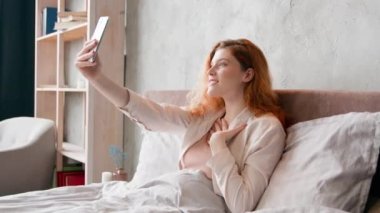 Mutlu, beyaz bir kadın yatakta cep telefonu uygulaması ile görüntülü konuşma yapıyor. Merhaba diyor, gülümsüyor, internetteki konferansa gülüyor, kaygısız kız blogger vlogger sabah yatak odasında video kaydı yapıyor.