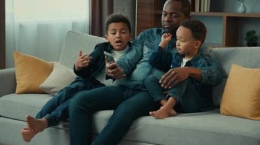 Afro-Amerikan mutlu bir aile evde cep telefonu uygulaması kullanarak akıllı telefon servisiyle eğleniyor. İki oğlu olan baba kanepeye oturmuş cep telefonlarını karıştırıyor fotoğraf maskesi deniyor.
