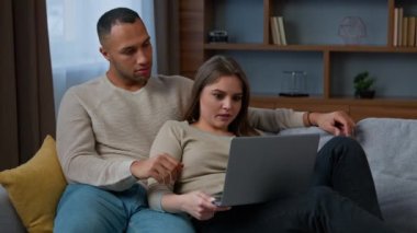Çevrimiçi alışveriş yapan çok ırklı çift ev sahipleri ev sahibi karı-koca sohbetlerinde dizüstü bilgisayarlı kanepeye sarılıyorlar. Emlak sitesinde daire seçip internetten internet ürünleri alıyorlar.
