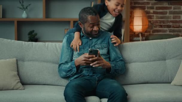 アフリカ系アメリカ人男性お父さん使用携帯電話ブラウジングスマートフォンでホームソファソファ2小さな子供息子男の子突然登場抱擁巨大パパ幸せな家族ボンディング父親 — ストック動画