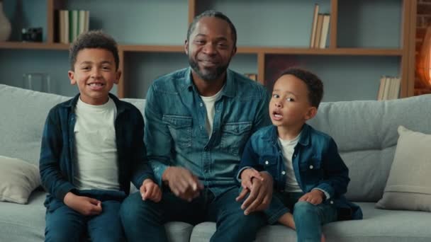 幸せな家族アフリカ系アメリカ人の父と兄弟少年たち子供は家でテレビを見ますテレビ番組お父さんPointing Toカメラお父さんとともに息子の肖像画でリビングルーム笑顔ウェブカメラビデオ通話 — ストック動画