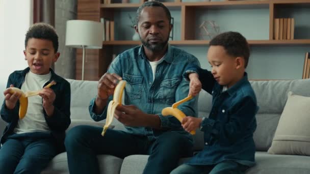 非裔美国父亲爸爸爸爸教两个孩子教小男孩儿子剥皮香蕉表演解释如何剥皮香蕉水果健康营养发展快乐家庭在沙发上的家 — 图库视频影像